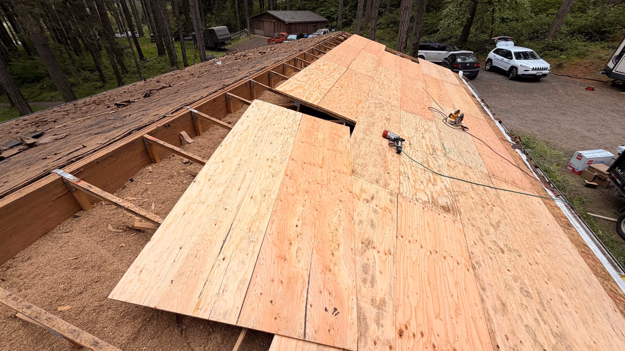 Partially re-roofed structure with new plywood sheets installed next to exposed wooden beams and old roofing materials in a wooded area with cars parked nearby.