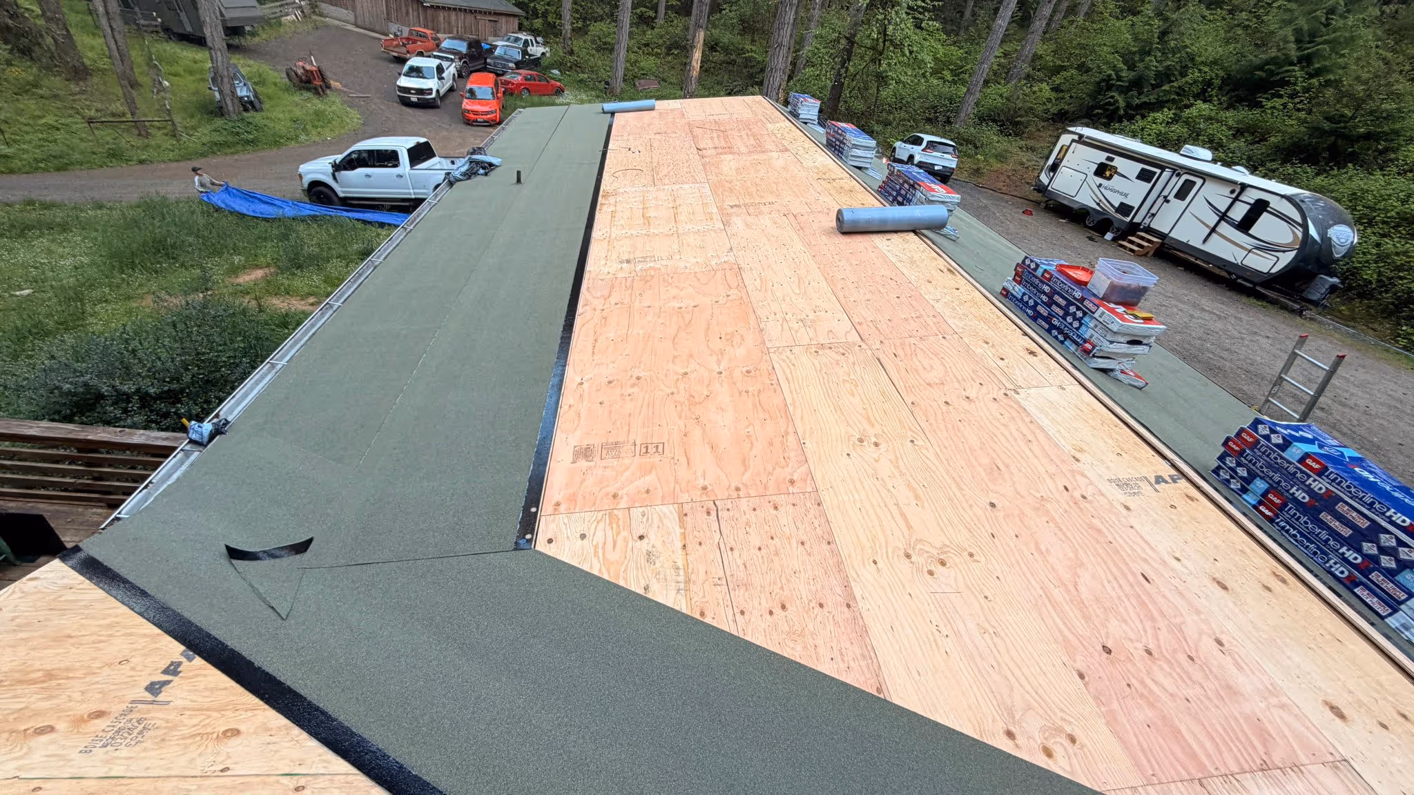 Partially shingled roof with exposed wooden plywood and roofing materials stacked on the side, surrounded by trees and vehicles.