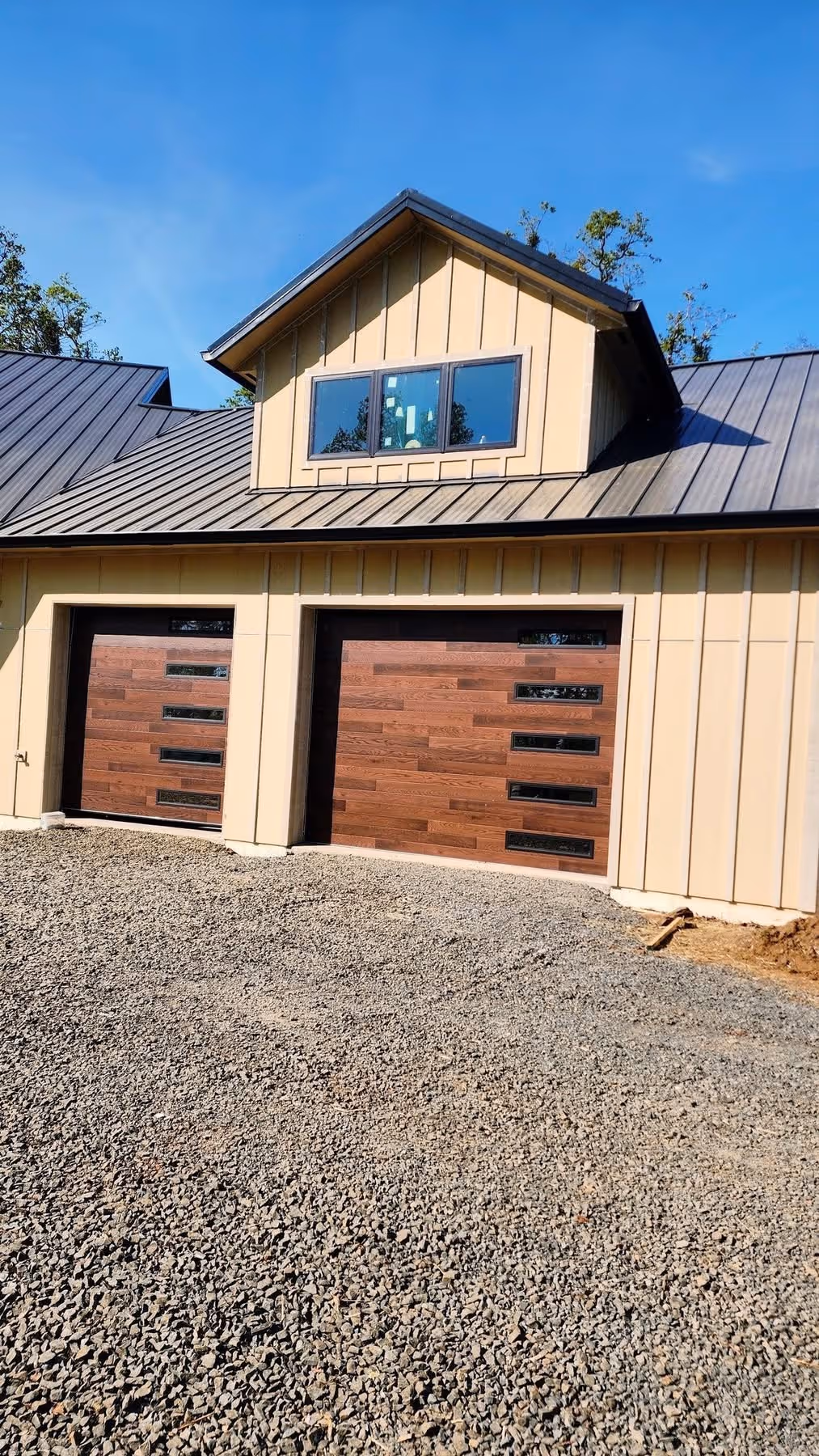 Beige house with black metal roof, featuring two wooden garage doors with horizontal windows and a dormer window above.
