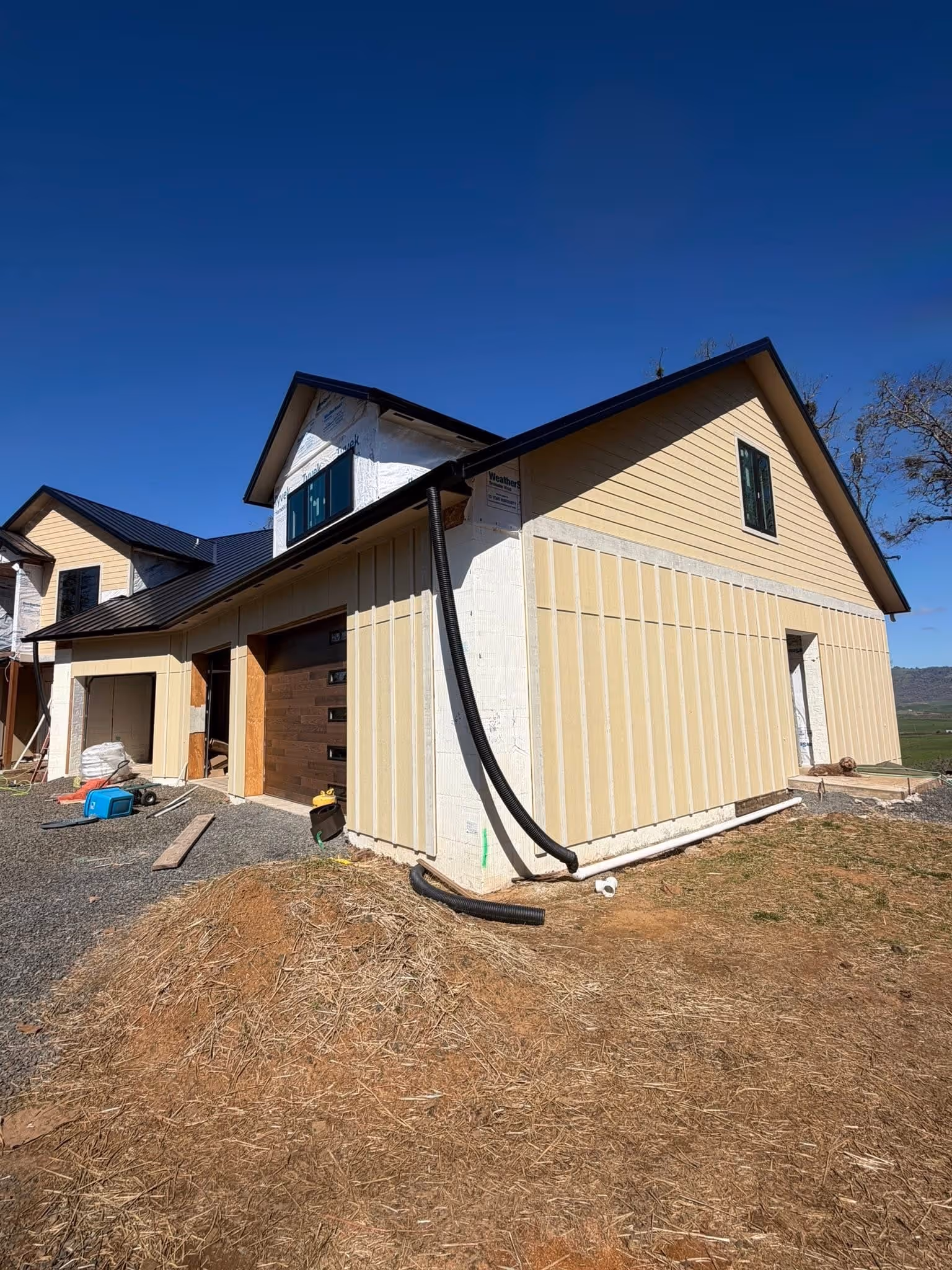 Partially constructed two-story house with beige siding, black metal roof, and construction materials on gravel and dirt ground under clear blue sky.