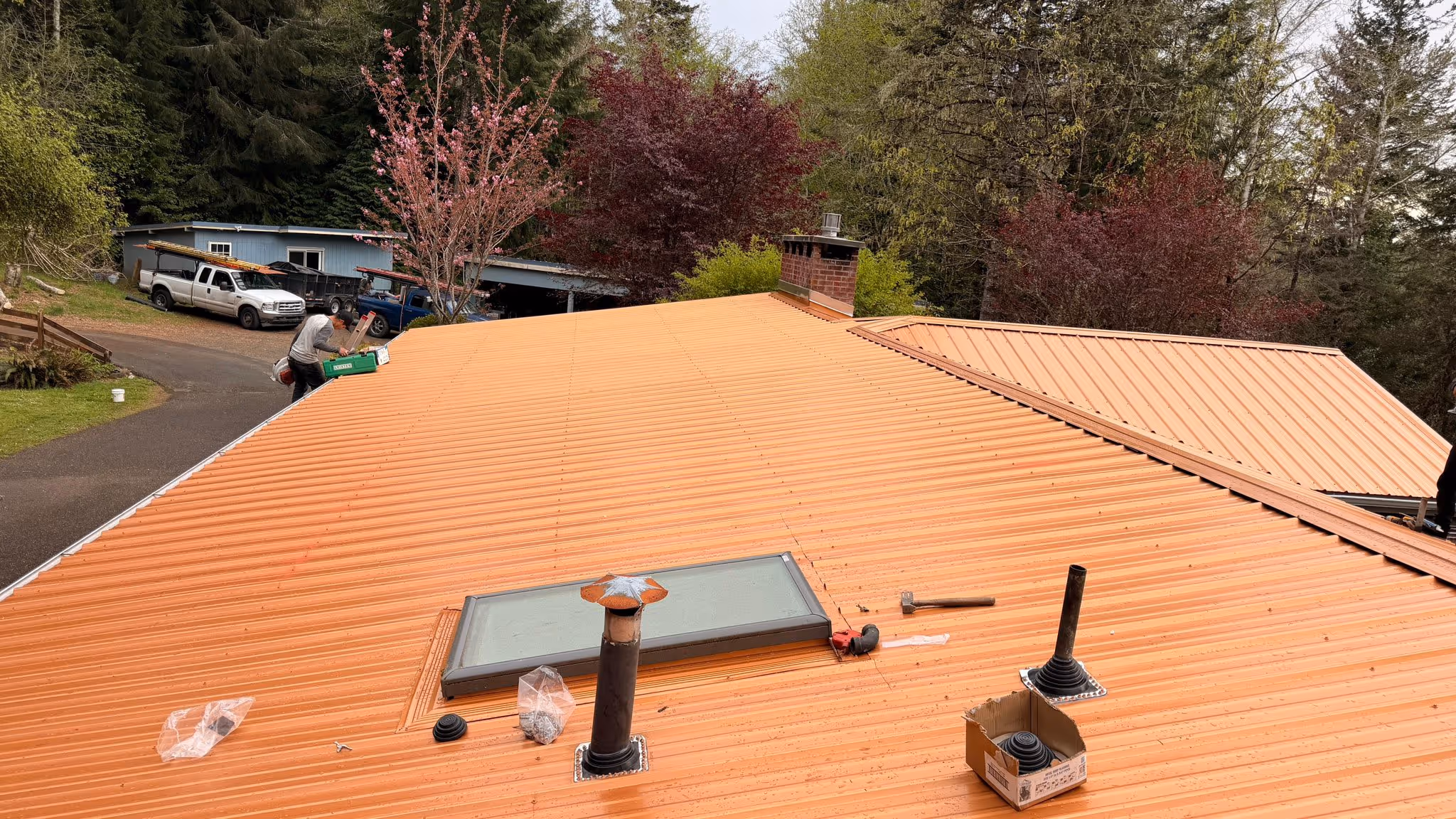 Newly installed orange metal roof with a skylight, chimney, and two ventilation pipes amid trees and parked trucks.