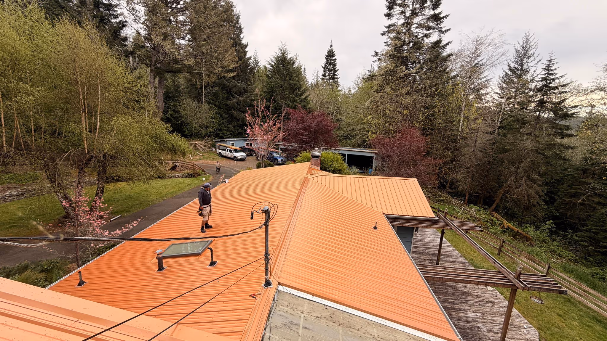 Person standing on a large orange metal roof of a house surrounded by trees and a driveway with parked vehicles.