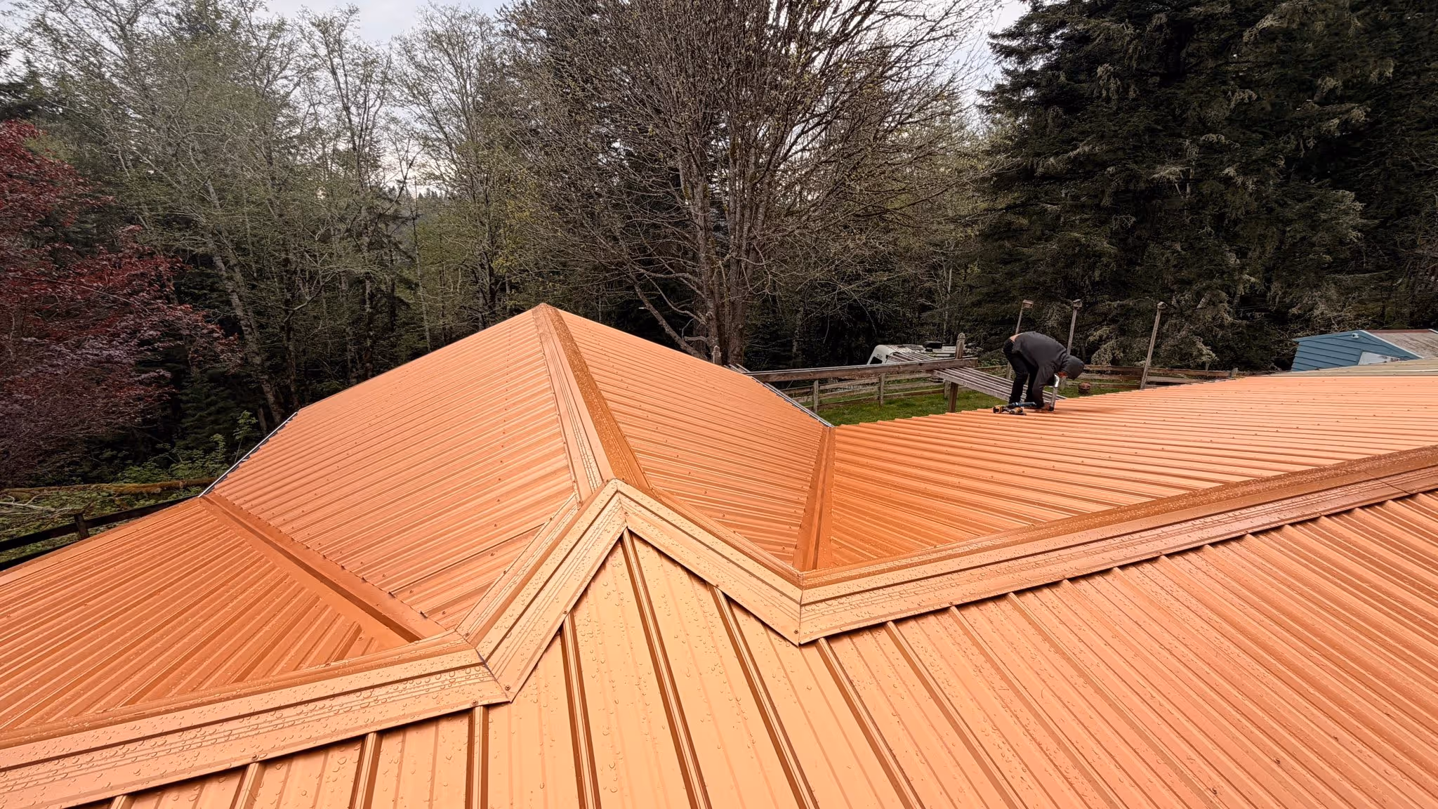 Worker installing an orange metal roof on a large building surrounded by trees.