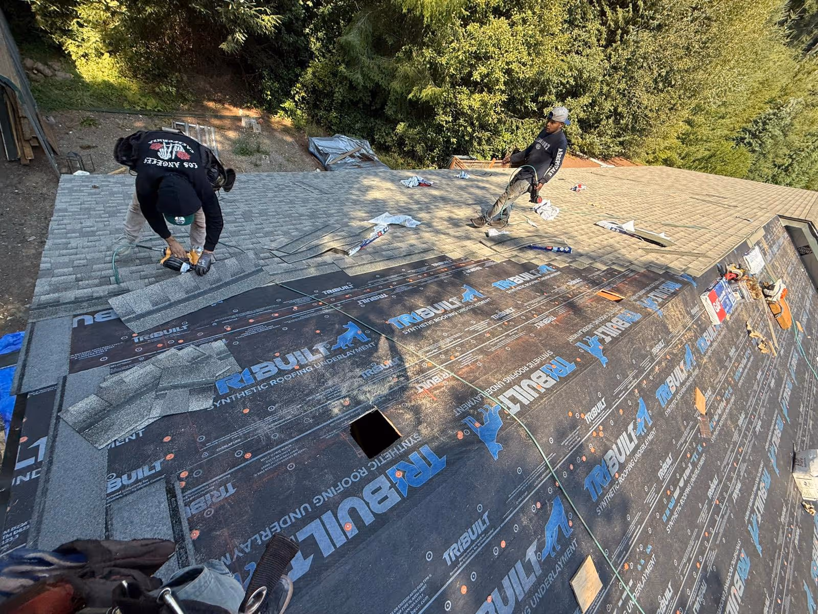 Two workers installing gray asphalt shingles on a roof with synthetic roofing underlayment labeled Tribuilt.