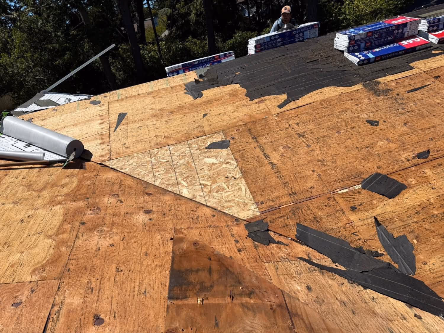 A roof under repair showing exposed plywood sheathing and partially removed old roofing material with roofing shingles stacked on the side.
