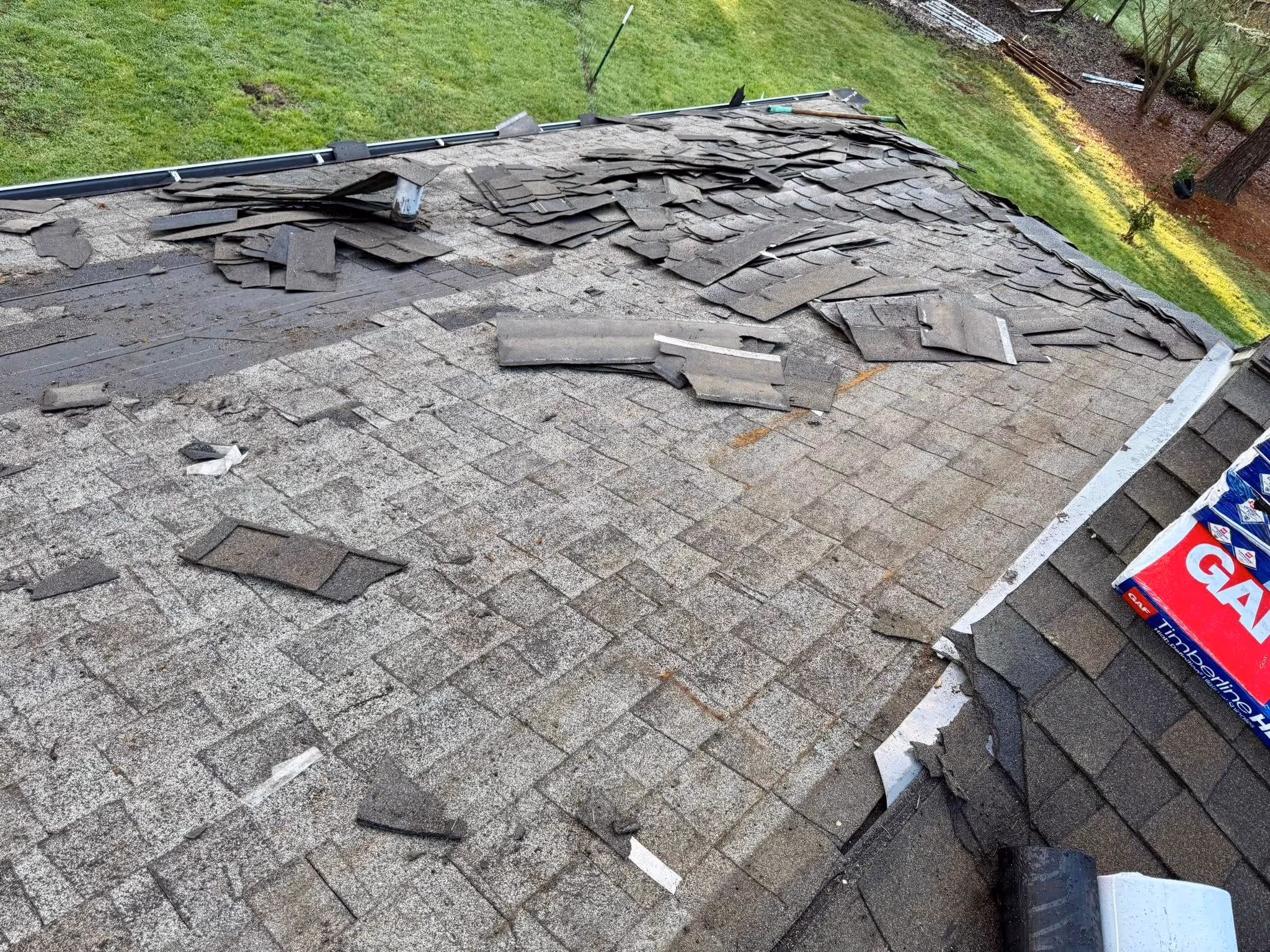 Roof with damaged and partially removed shingles next to a green lawn and trees.