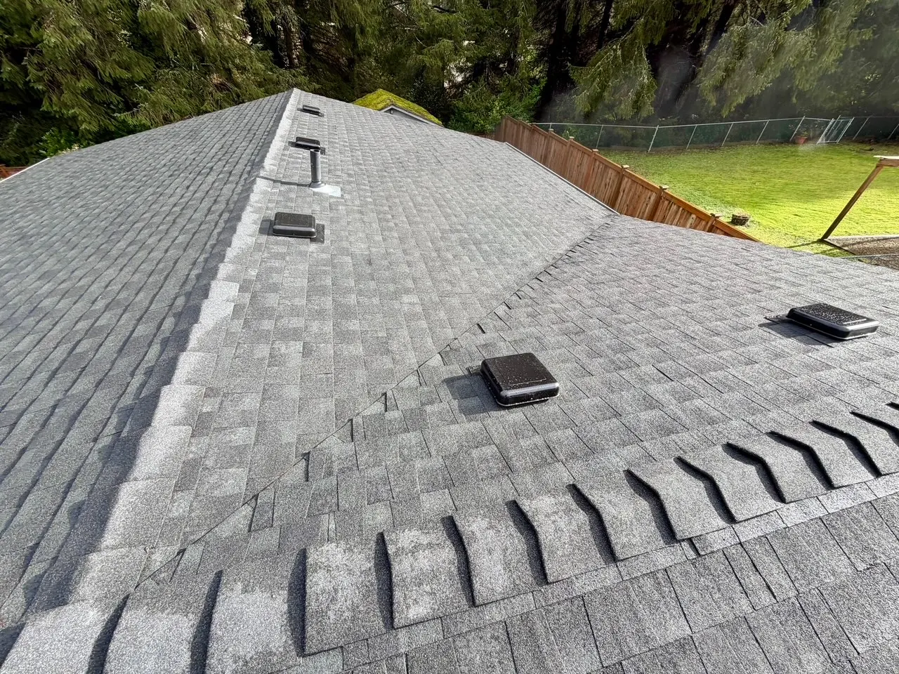 Gray asphalt shingle roof with multiple black vents and surrounded by trees and a wooden fence.