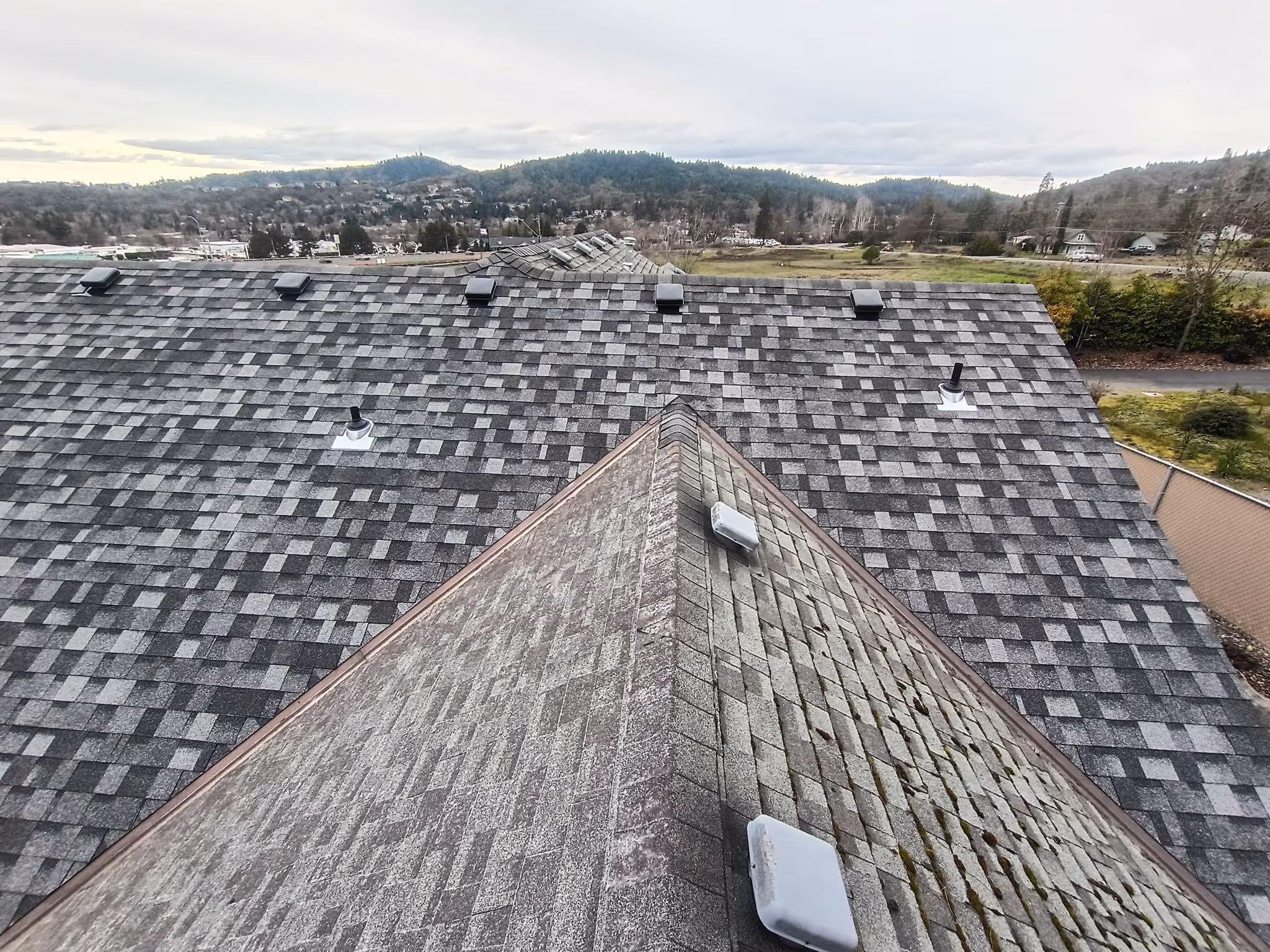 View of gray shingled rooftops with ventilation pipes and vents, set against a background of hills and cloudy sky.