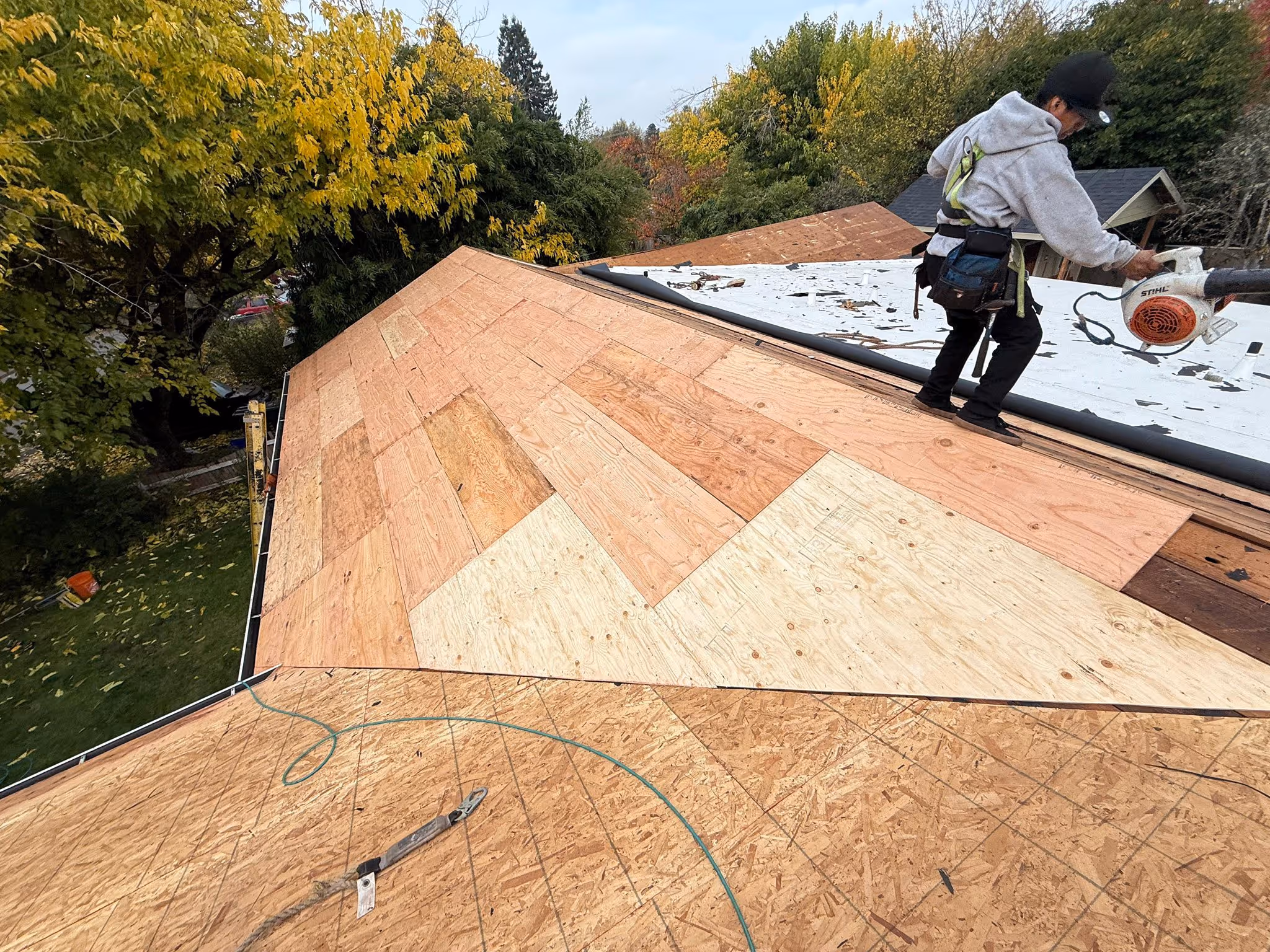 Worker wearing safety gear standing on a partially plywood-covered roof using a leaf blower, surrounded by trees with autumn foliage.