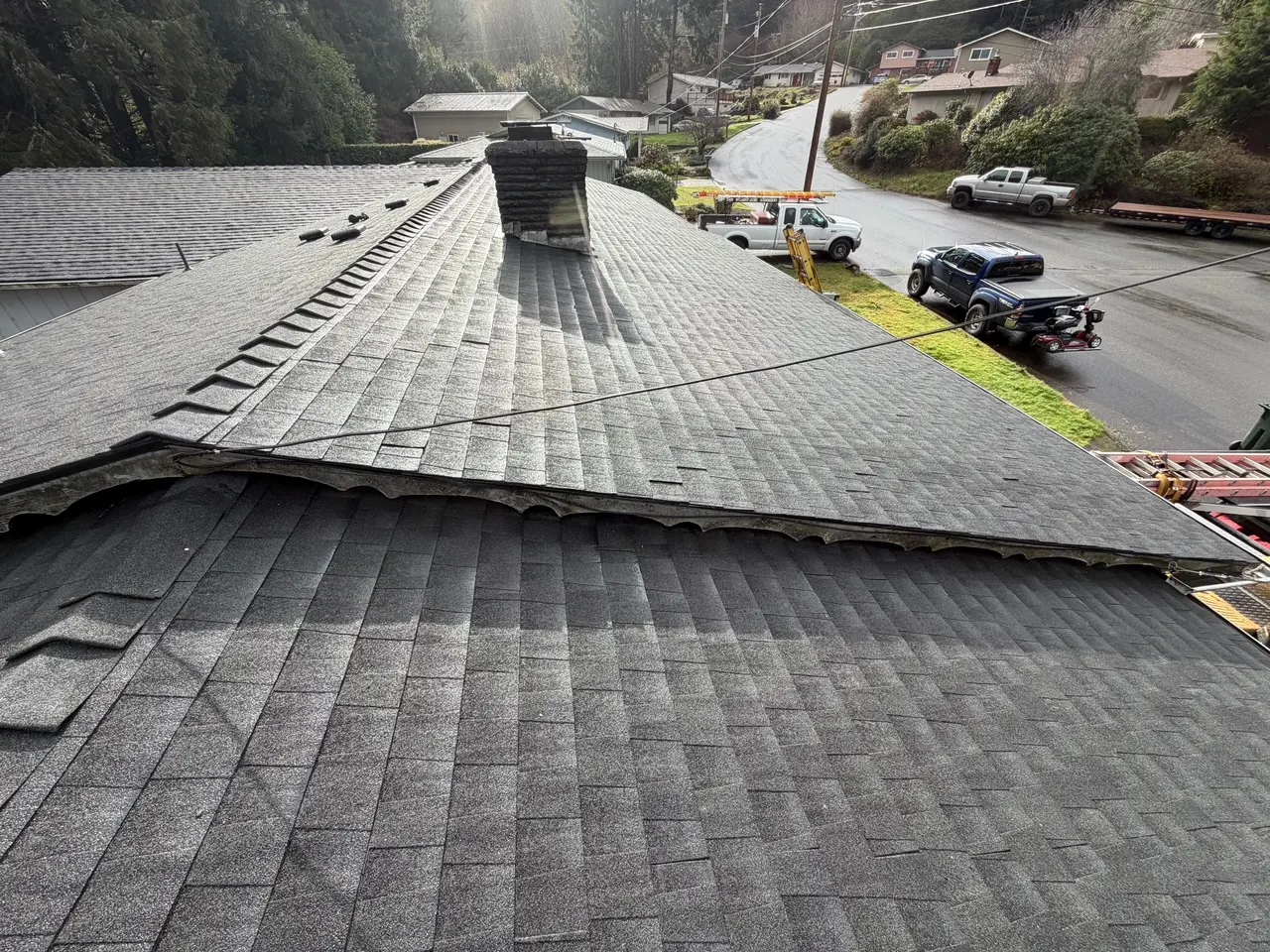 New grey asphalt shingle roof on a house with a chimney, surrounded by trees and several parked vehicles on the street.