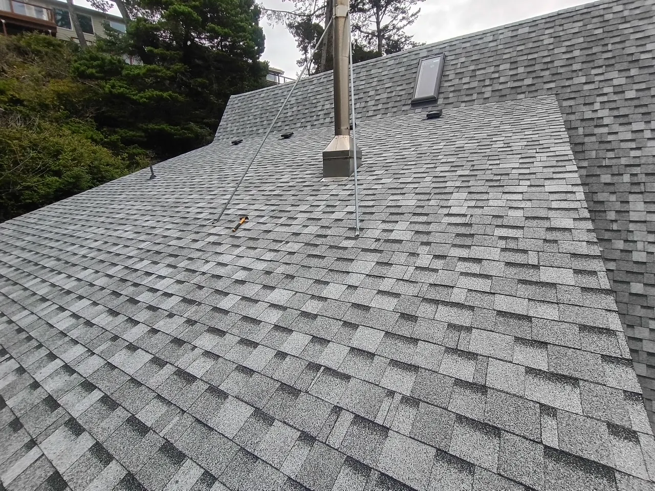 Grey asphalt shingle roof with a metal chimney and a small skylight, surrounded by trees.