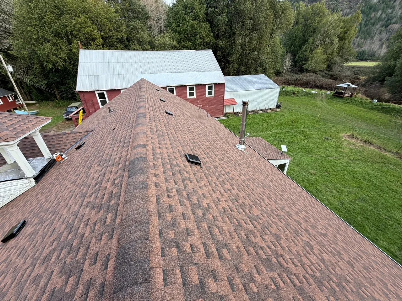 Bird's-eye view of a brown shingle roof with vents and a chimney, overlooking green lawn and trees with a red barn and a metal shed in the background.