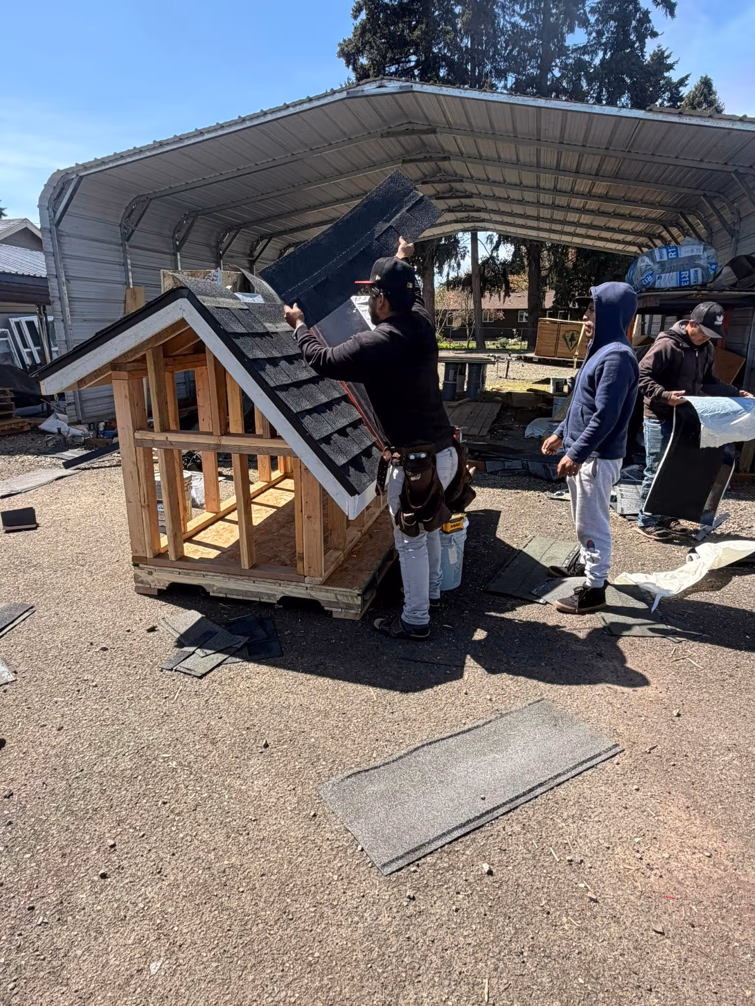 Three men working outside on constructing a small wooden structure with a black shingled roof under a metal carport.